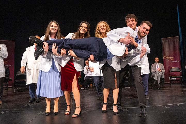 Four medical students stand on a stage holding a male student across their arms as they smile for the camera.