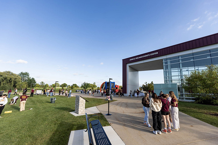 A small group of students stand outside the CMU Events Center as other groups of students stand in the distance during a football tailgate party.