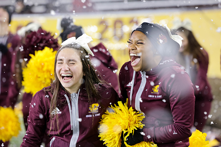 Two cheerleaders wearing CMU team jackets laugh and smile during a snow storm at the CMU football game.