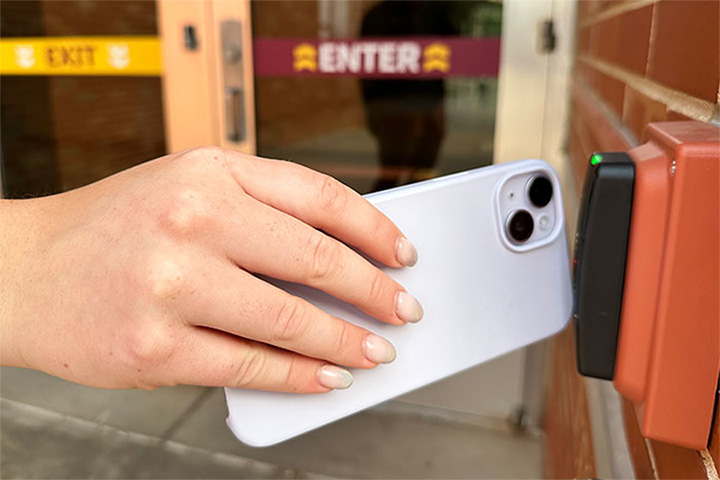 A hand holds a mobile phone in a white case near an orange and black digital door access reader.