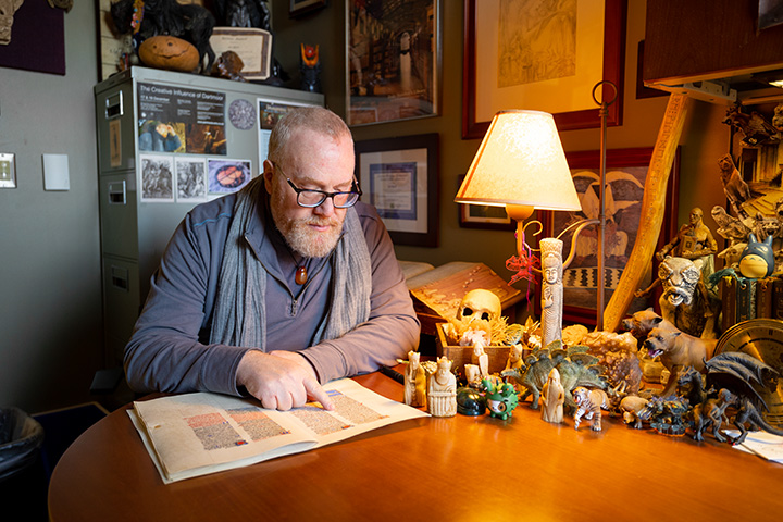 A male professor wearing glasses reads a book at a desk covered with figurines and small statues.