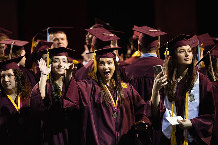 A group of CMU graduates in maroon robes and hats smiles and wave at the camera.