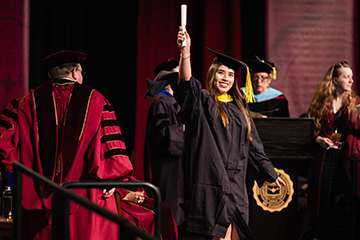 A CMU graduate stands on stage holding her diploma high over her head while smiling.