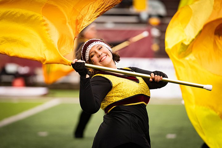A color guard member wearing black and gold smiles at the camera while waving a large yellow flag.