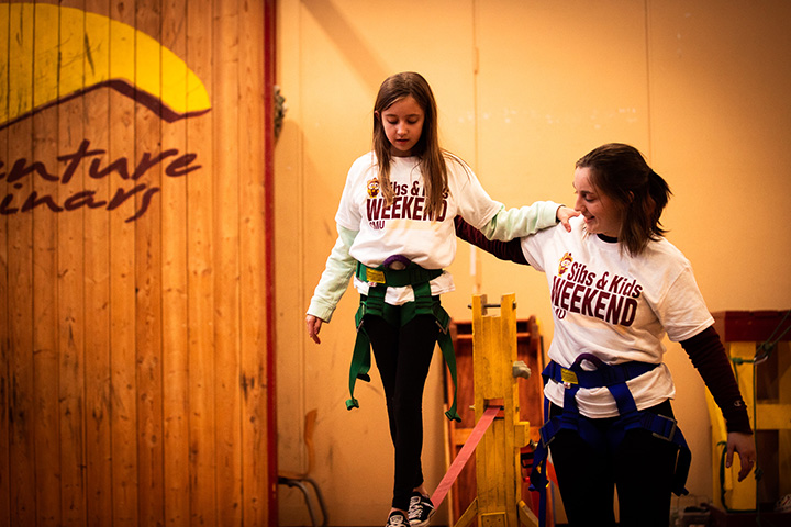 A child walks on a tight rope three feet above the ground as a CMU student walks alongside her for support.