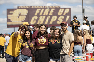 A group of students wearing CMU apparel smiles at the camera during a football pregame tailgate event as a giant Fire Up Chips! sign stands tall in the background.