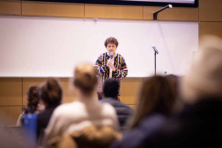 A woman in a colorful sweater stands on stage holding a microphone while talking about her experience as a Holocaust survivor.