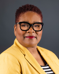 A woman wearing a yellow jacket and glasses smiles at the camera.