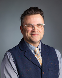 A man wearing a vest, tie and glasses smiles at the camera.