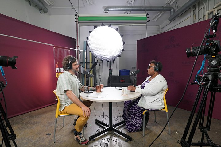 Adam Sparkes and Nikita Murry wear headphones and sit at a round table in a studio. Bright lights shine own with maroon backdrops behind them and cameras on tripods flanking them on either side.