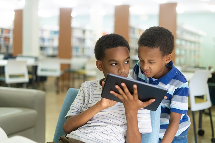A boy in a white and blue striped shirt holds a tablet in his hand while another boy with a blue striped collared shirt looks over his shoulder.