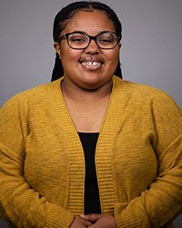 Professional headshot of Michyah Jones in a gold cardigan over a black top against a gray background.