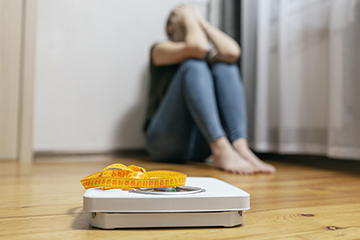 A woman sits against the wall sobbing with a tape measure on top of a scale in the foreground.