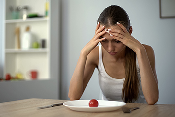 A thin woman with long hair wearing a white tank top looks at a white plate containing a single red cherry tomato.