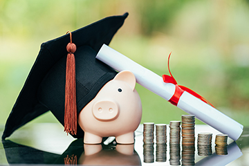 A black mortarboard sits atop a piggy bank next to stacks of coins with a diploma leaning against it.