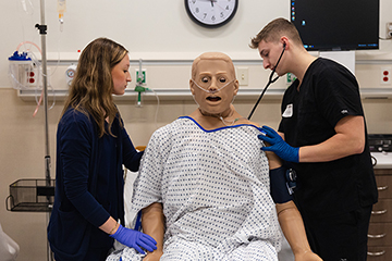 A male nursing student places a stethoscope on the back of a medical dummy while a female nursing student assists.
