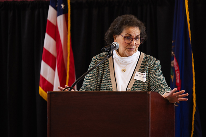 A woman in glasses wearing a white turtleneck and a sweater vest talks from behind a podium with an American flag in the background.