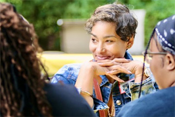 A smiling woman rests her chin on her folded hands while talking to two college students.