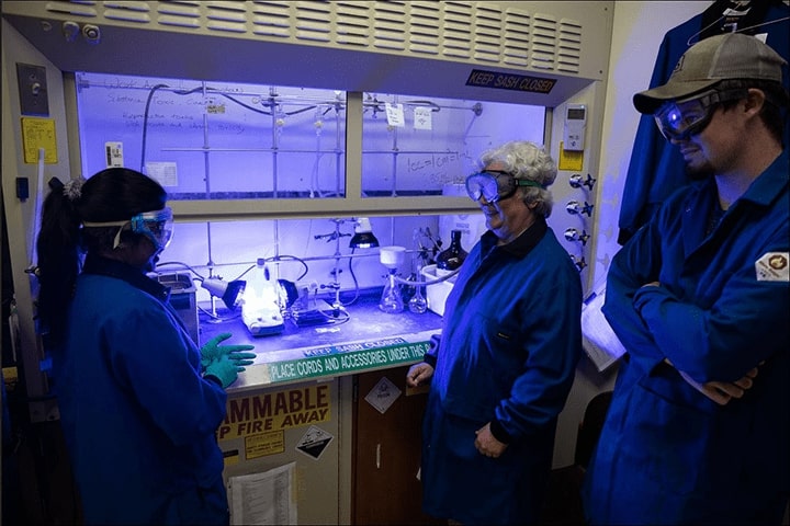 Three researchers wearing goggles and blue lab coats stand in front of a fume hood lit by blueish LED lights and with stickers that say Keep sash closed, Place cords and accessories under this and Flammable keep fire away.