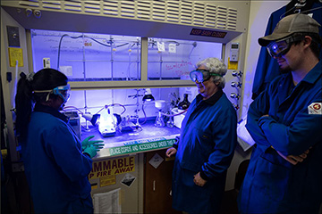 Three researchers wearing goggles and blue lab coats stand in front of a fume hood lit by blueish LED lights and with stickers that say Keep sash closed, Place cords and accessories under this and Flammable keep fire away.