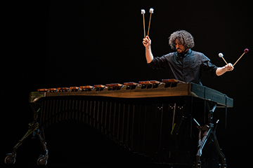 A man dressed in black with shaggy medium-length black hair hits a marimba, a musical instrument that looks like a xylophone, with two mallets.