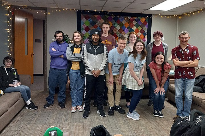 Ten smiling young people of mixed race and gender and wearing an assortment of clothing stand in the center to the right side with an eleventh sitting on a couch on the left side.
