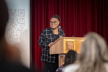 A woman with glasses and dressed in a dark suit smiles while standing behind a podium against a background of red drapery.