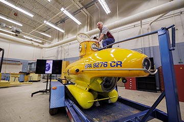 A silver-haired man in blue jeans and a gray and maroon polo shirt partially stands atop a yellow miniature submersible that is on a blue trailer in the middle of a laboratory.
