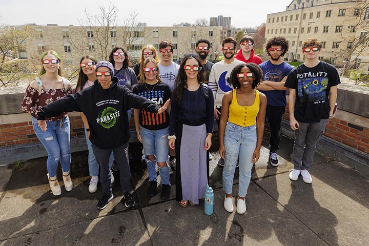 A group of people wearing solar eclipse glasses pose together while standing on a roof.