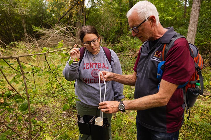 A man in a short-sleeved shirt and wearing glasses and a backpack pulls a sample from a test well while a woman in glasses and brown hair, and wearing a gray sweatshirt, watches.