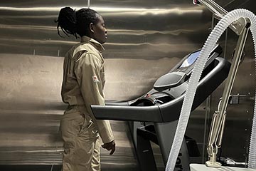 A woman with black hair pulled back and wearing a beige jumpsuit walks on a treadmill against a metallic background.