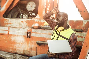 A man in a checkered shirt and yellow safety vest holding a laptop wipes his forehead with his other hand under a hot sun.