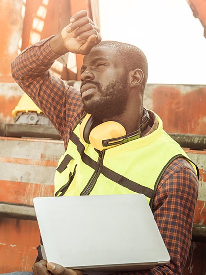 A man in a checkered shirt and yellow safety vest holding a laptop wipes his forehead with his other hand under a hot sun.