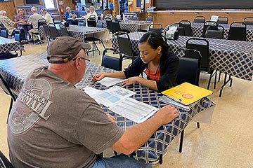 A woman with parted black hair wearing a black sweater over a red top fills out paperwork with a man in a T-shirt and a ball cap at a folding table with a diamond pattern.
