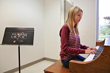 A woman with long blonde hair wearing a red and white striped top and blue jeans sits at a piano with a music stand behind her on which are written the words, School of Music and CMU.