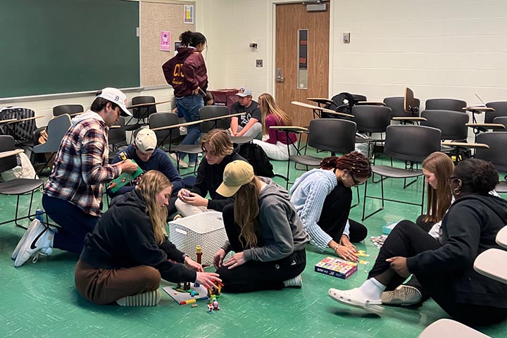 Four groups of young people, three sitting on the floor, are gathered in a classroom.