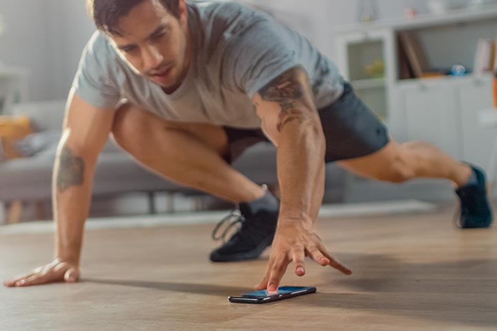 An athletic man with brown hair on his head and face and wearing workout clothing scrolls on his phone during a workout.