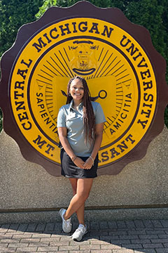A woman with long hair and wearing a gray shirt and dark shirt stands in front of a university seal of maroon and gold.
