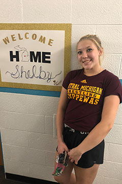A young woman with blonde hair pulled back smiles while standing next to a sign that reads, Welcome Home Shelby.
