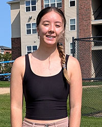 A young woman in a black top and peach-colored pants smiles while standing outside.