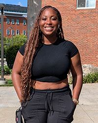 A young, smiling woman wearing a black top and pants standing outside.