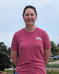 A young, smiling woman wearing a pink T-shirt standing outside.
