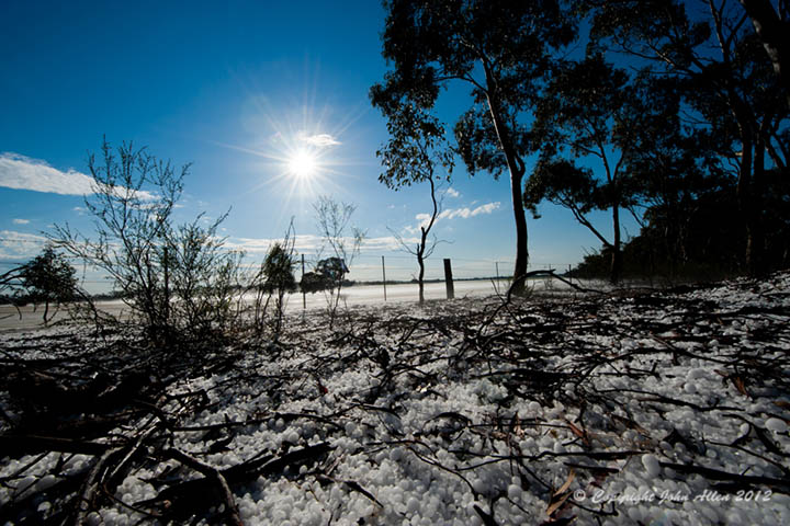 Hail stones cover a farm field under a sunny sky.
