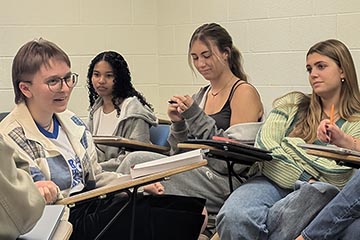 A young woman sitting in a classroom desk with short hair and glasses speaks while three other young women, also seated in classroom desks, look on.