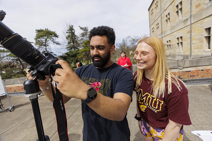 A man with curly dark hair and a beard and a woman with straight blonde hair and glasses look at a viewing screen attached to a camera.