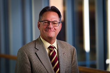 Tom Masterson wears a suit, yellow shirt and maroon and yellow tie while posing for a headshot photo.