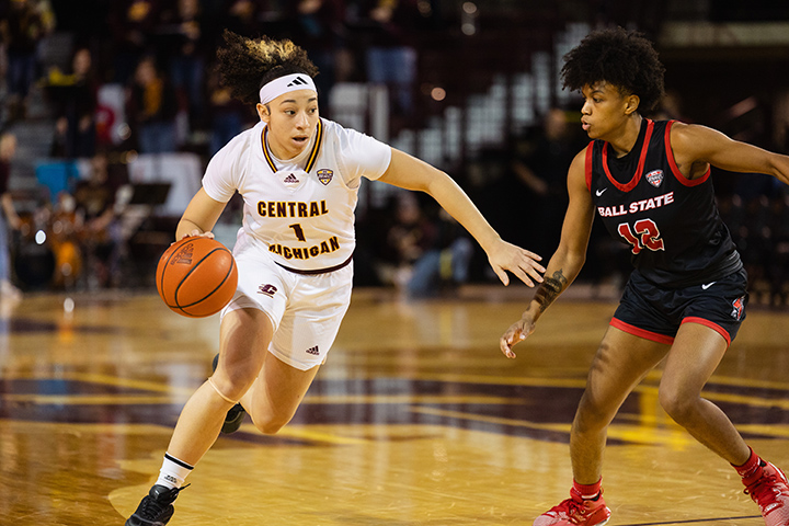 CMU women's basketball player Lisa Tesson wears a white uniform and headband, dribbling past a defender wearing a black and red uniform.