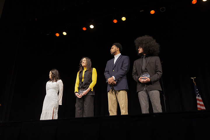 Four college students stand in a line on a stage facing the crowd after winning awards.