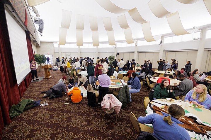 A large group of people sit at desks and on the floor inside the UC Rotunda room. The room is well lit with bright light shining from the ceiling.