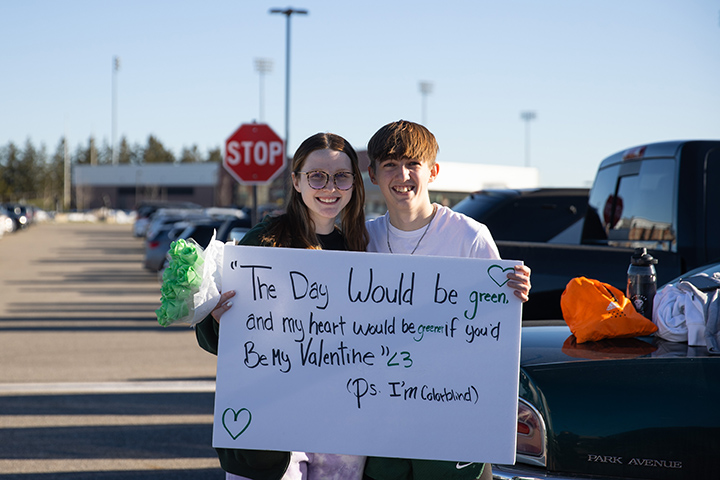 A male and female student stand in a parking lot holding green roses and a white poster board that reads 
